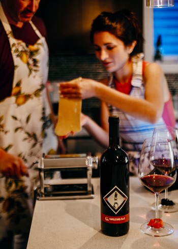 People make pasta in a kitchen with a bottle of red wine. 